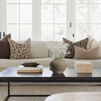 A styled living room scene featuring a light beige sofa decorated with brown, beige, and black patterned throw pillows, set behind a dark wood coffee table on a textured rug.