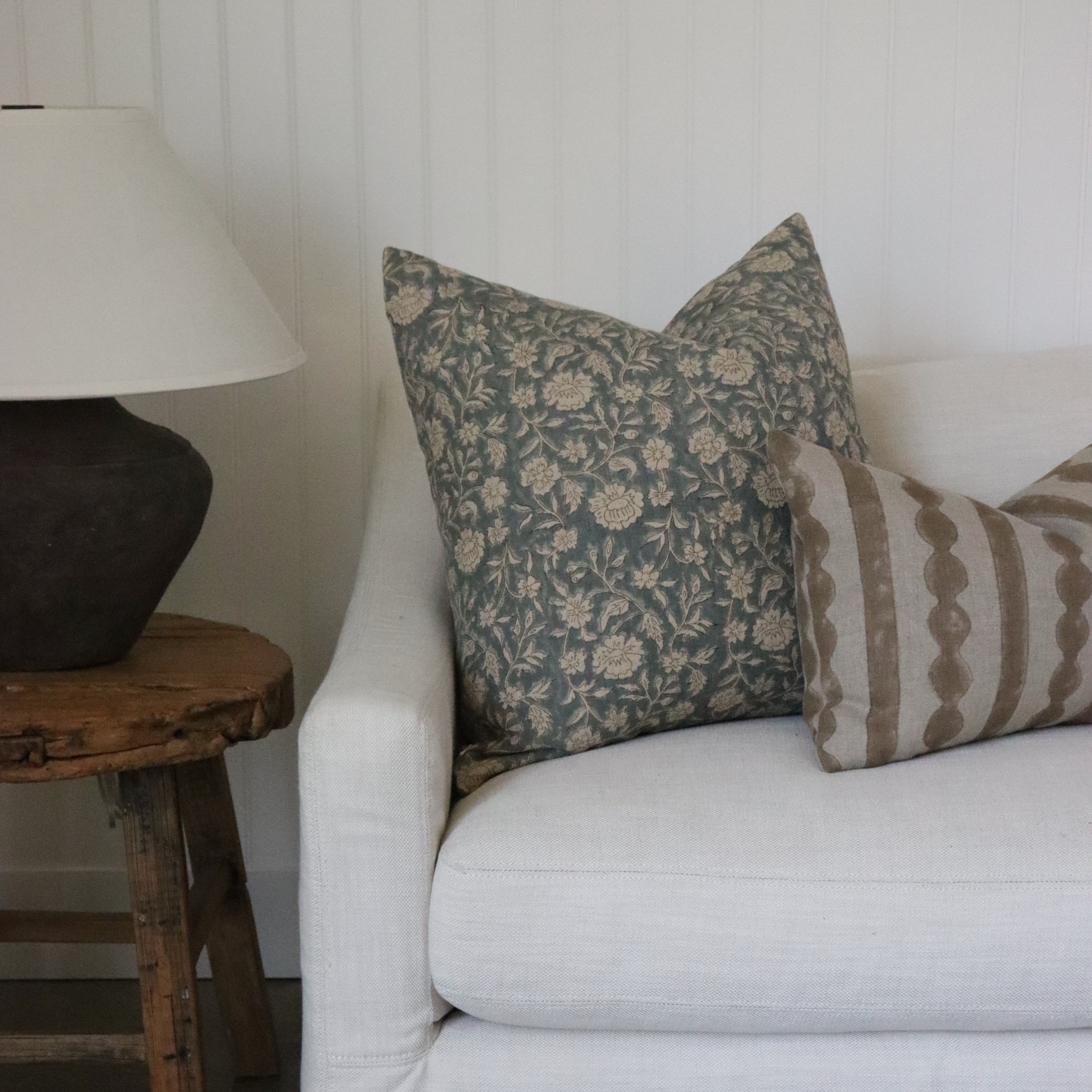 White sofa with patterned pillows against a white wall, wooden side table with lamp.